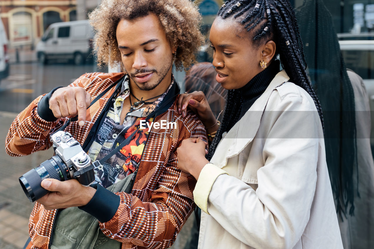 Young stylish african american male photographer with photo camera showing pictures to female model during session on city street