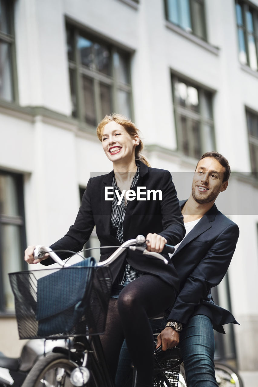 Happy businesswoman riding bicycle while colleague sitting on back seat outdoors