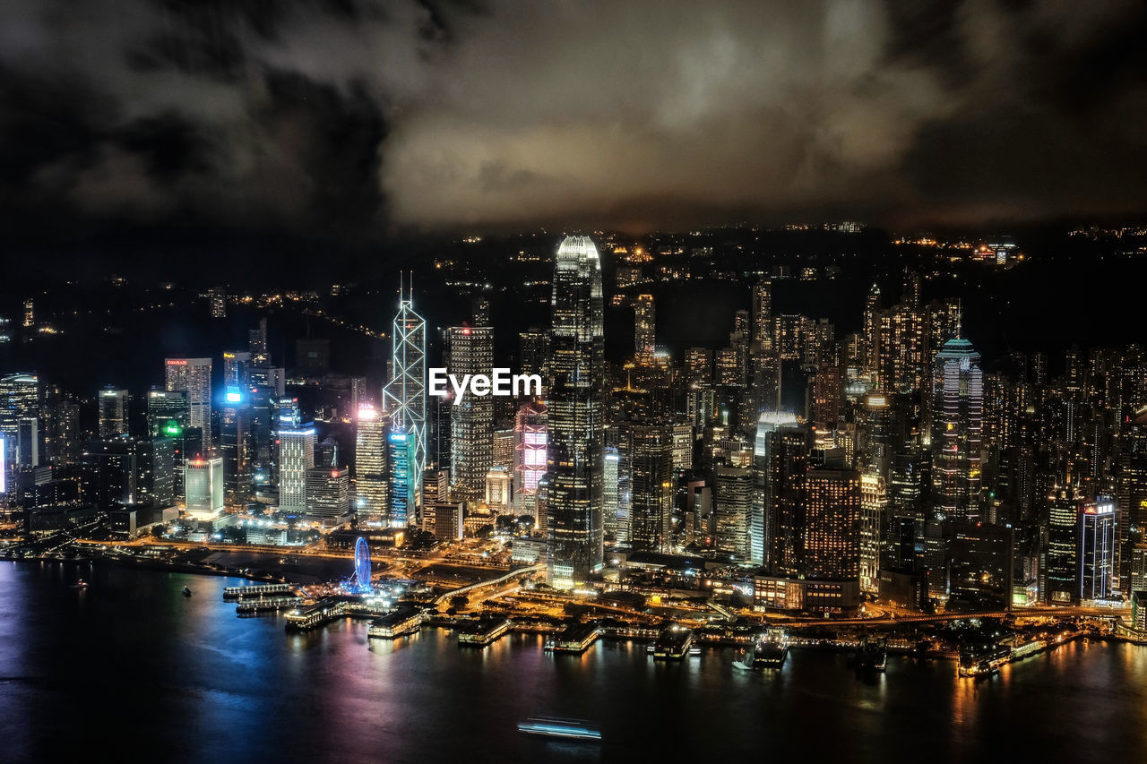 Illuminated city by sea against cloudy sky during night