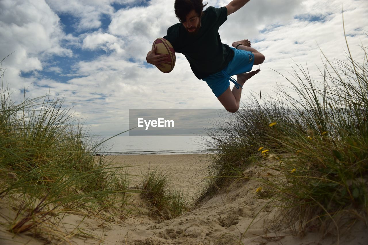 Man with american football jumping over grass at beach against sky