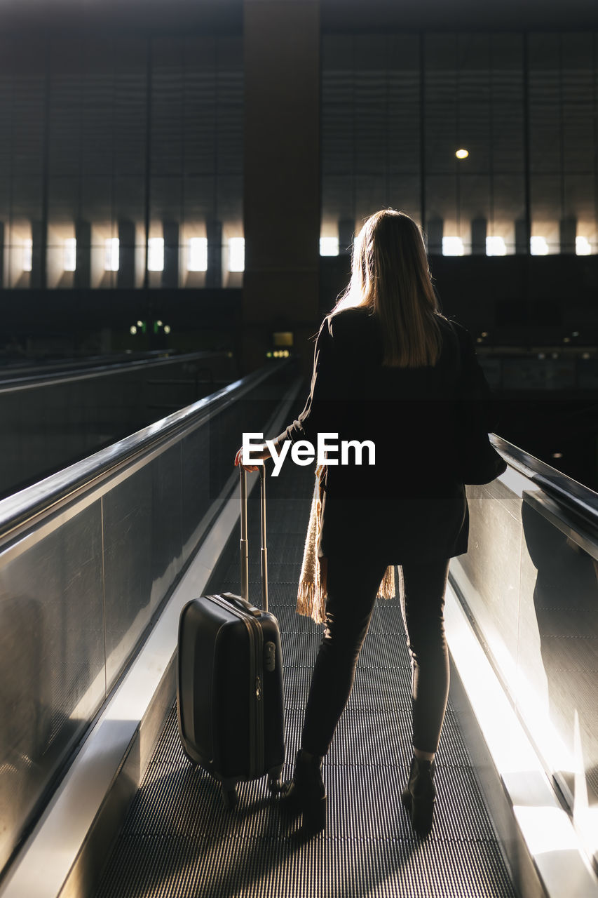 Woman with wheeled luggage moving on escalator at station