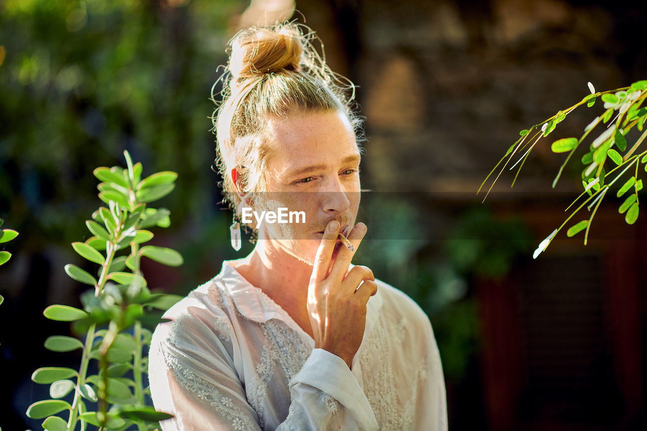 Thoughtful handsome blond male wearing hipster summer clothes looking away while standing smoking cigarette surrounded by green vegetation