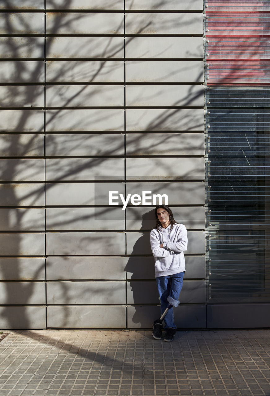 Young disabled man with arms crossed standing against wall with tree shadow