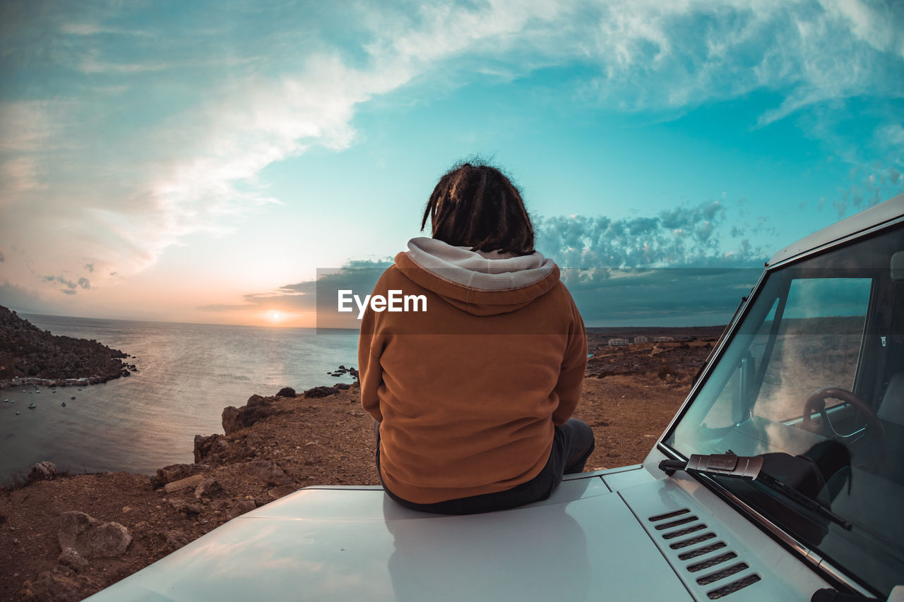 Rear view of young man with dreadlocks sitting by sea against sky during sunset