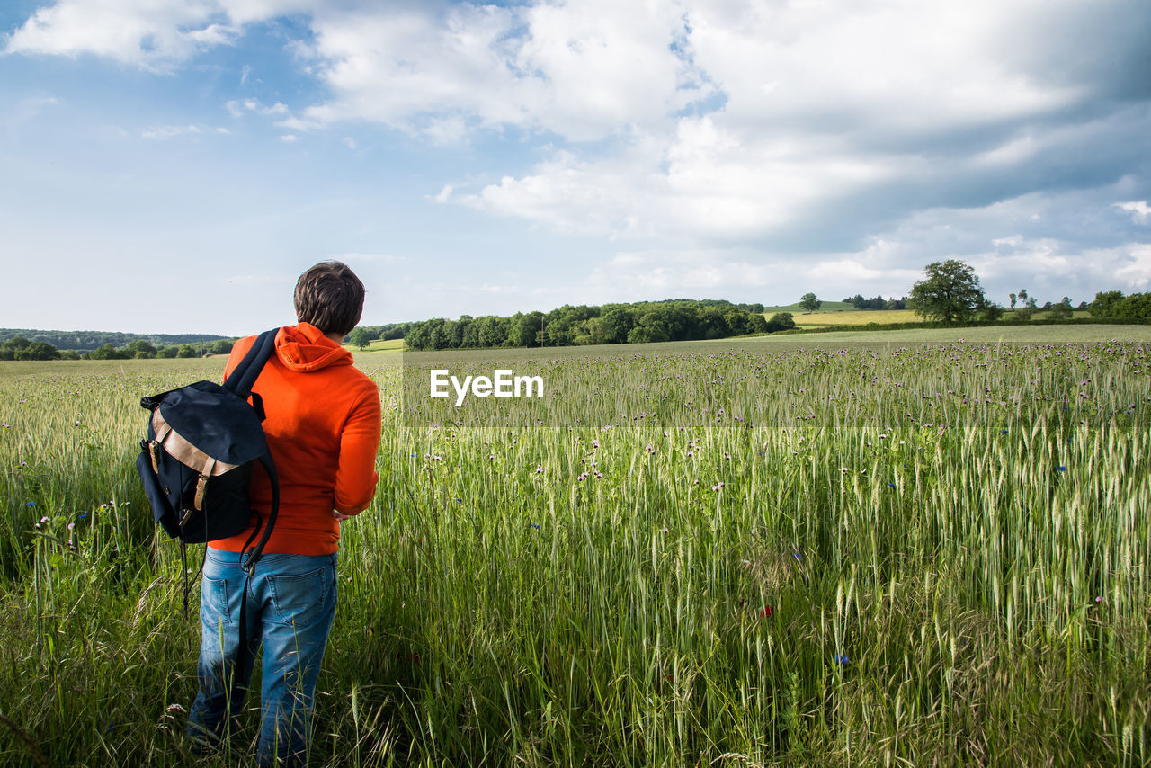 Rear view of backpack man standing on wheat field against cloudy sky