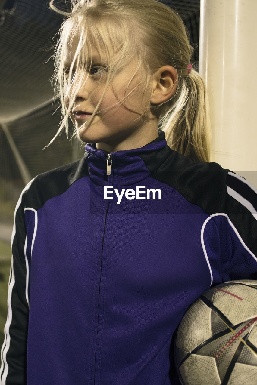 Girl holding soccer ball looking away while standing by goal post