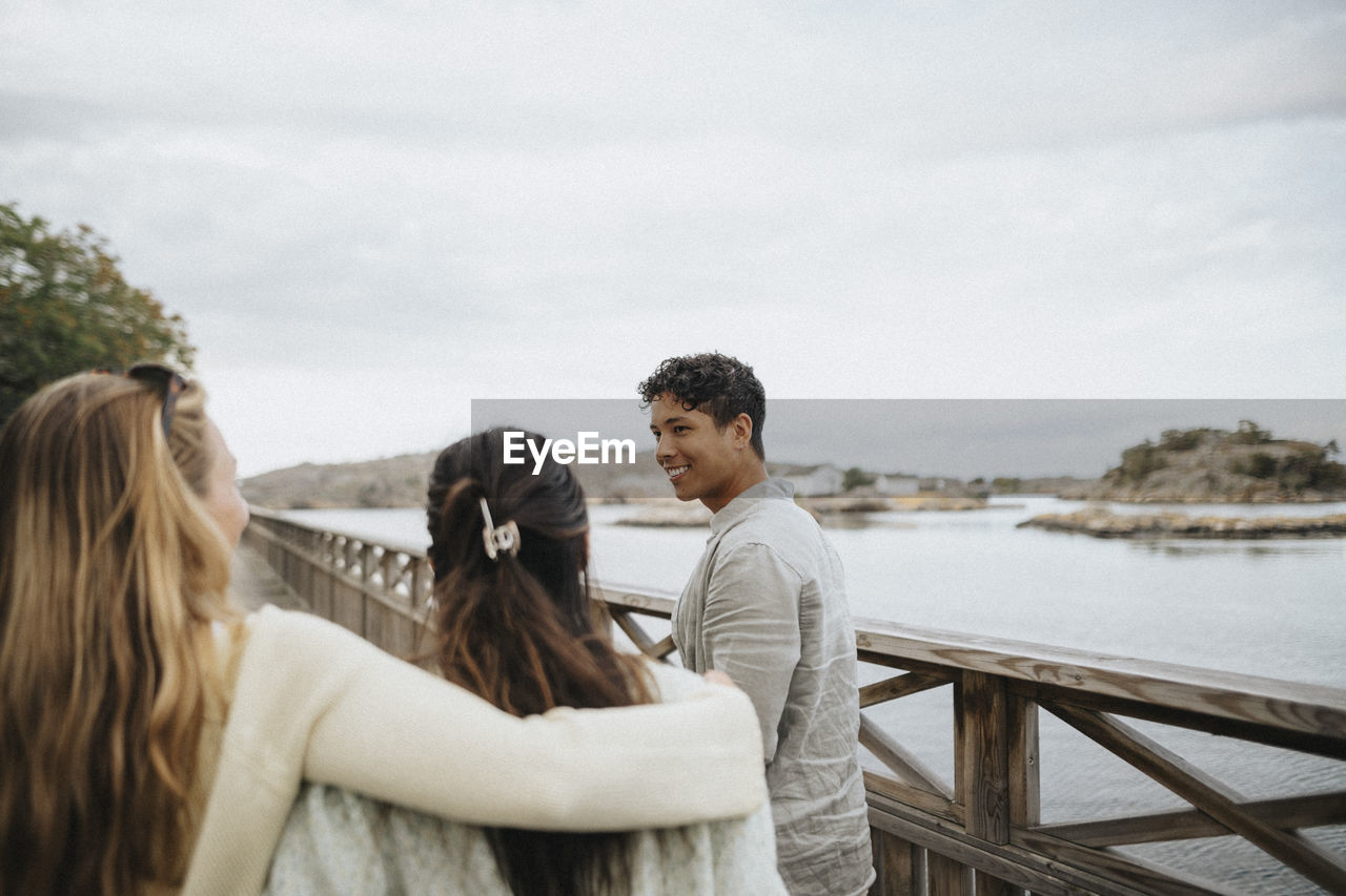 Smiling young man with female friends walking on bridge near lake