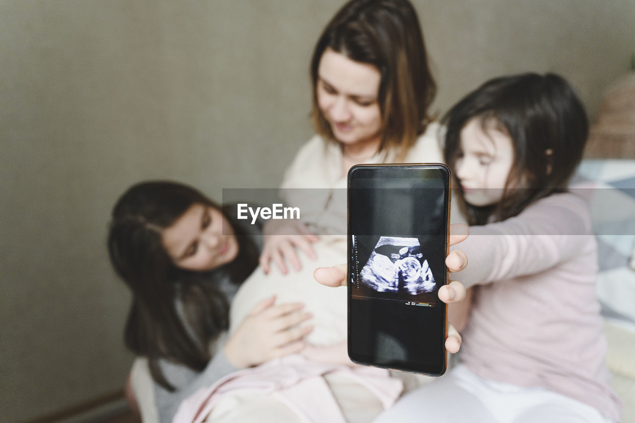Curious daughter with mobile phone showing ultrasound embryo of baby at home