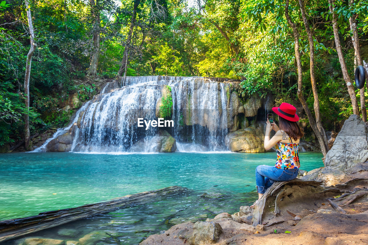 Side view of woman using mobile phone while sitting on rock against waterfall