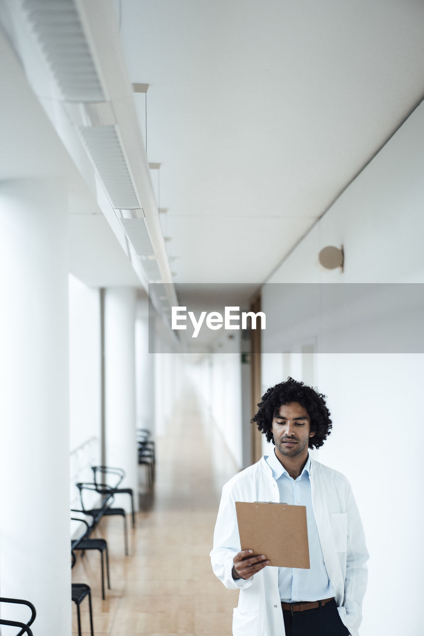 Confident young male doctor looking at clipboard in hospital corridor