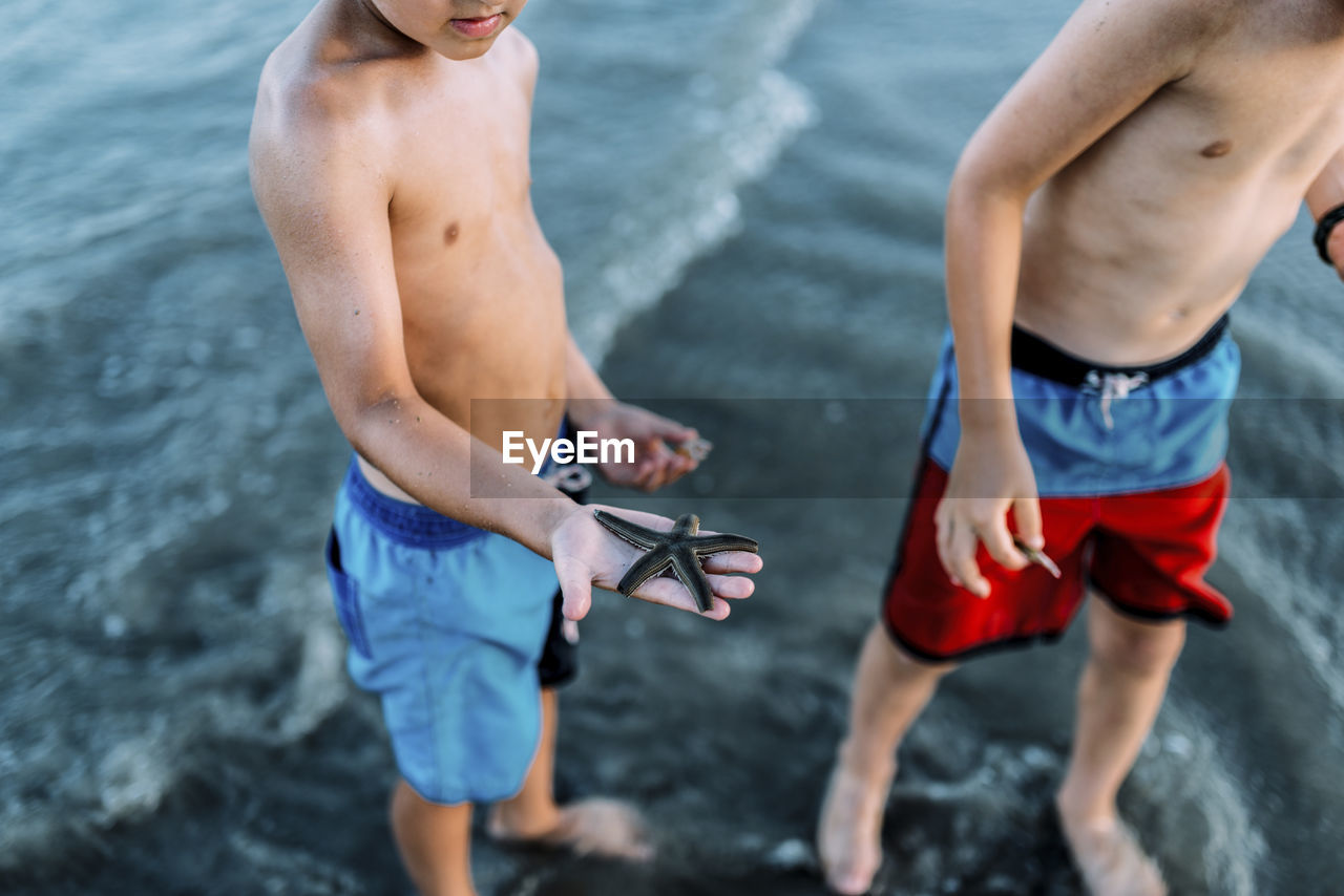 Low section of shirtless brothers standing at beach