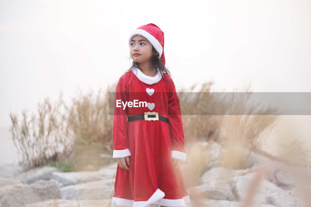 Cute girl wearing red dress while standing on rocks against sky during christmas