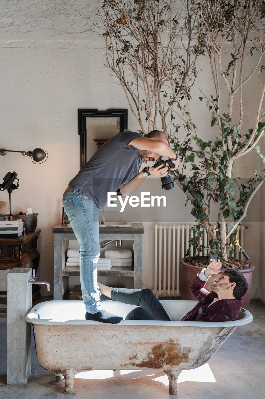 Side view dedicated man standing on bathtub edges and shooting from above adult guy in stylish clothes lying in aged white bath near big green potted tree against rustic vintage interior in photo studio