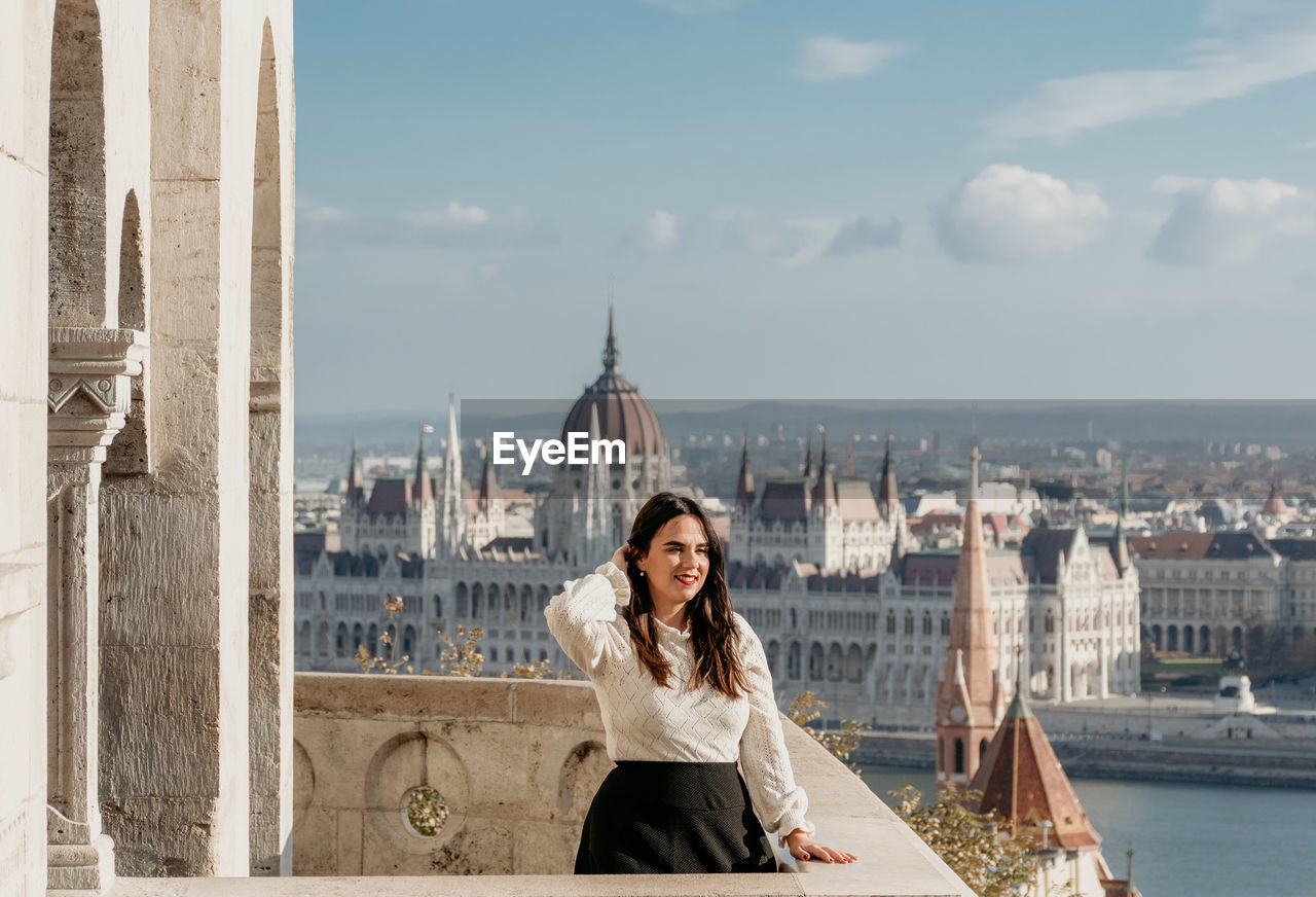 Portrait of beautiful young woman on balcony overlooking hungarian parliament in budapest, hungary