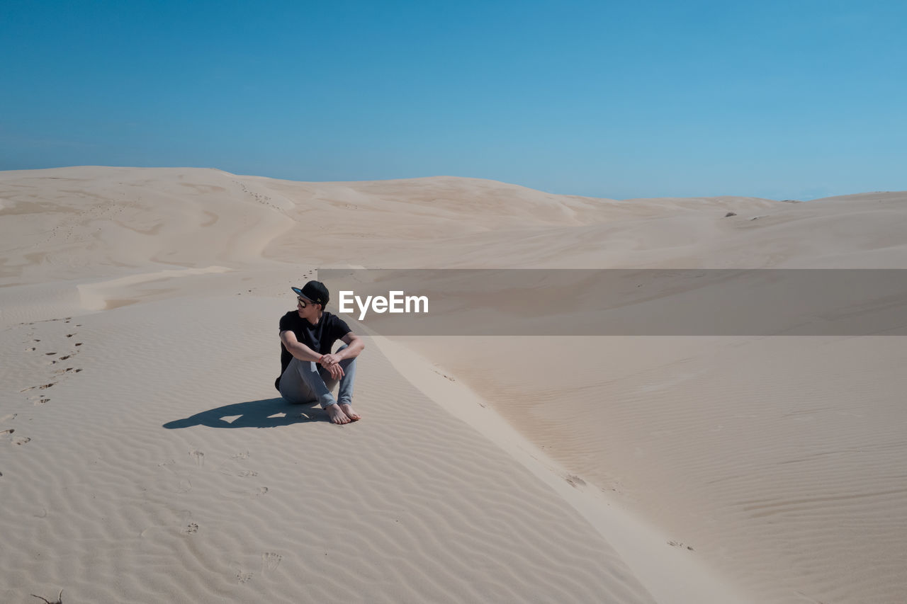 Full length of man sitting on sand in desert against sky