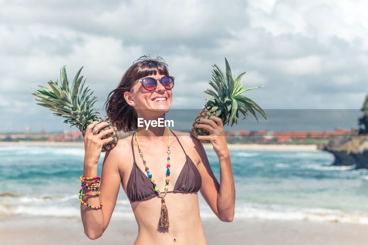 Happy young woman holding pineapples at beach against sky