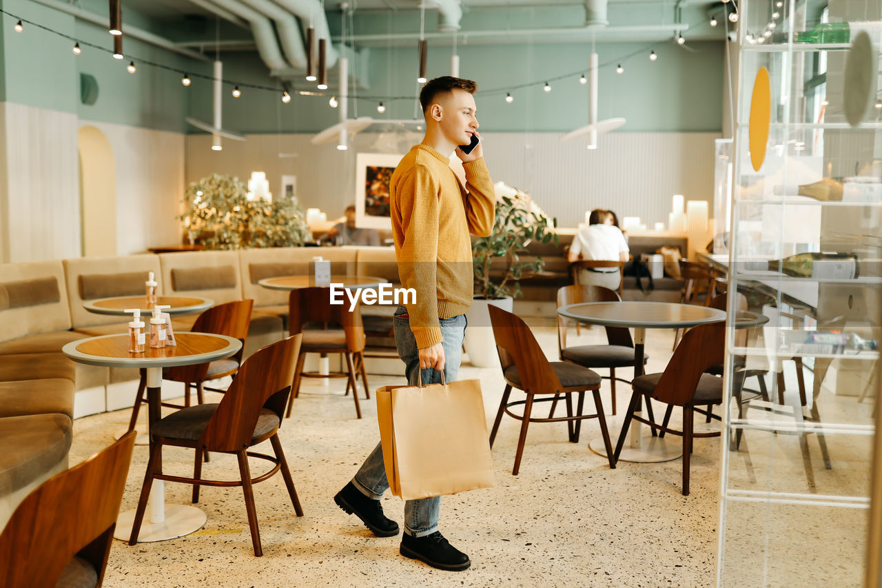 A young man in casual clothes with bags in his hands after shopping stands alone and talks in a cafe