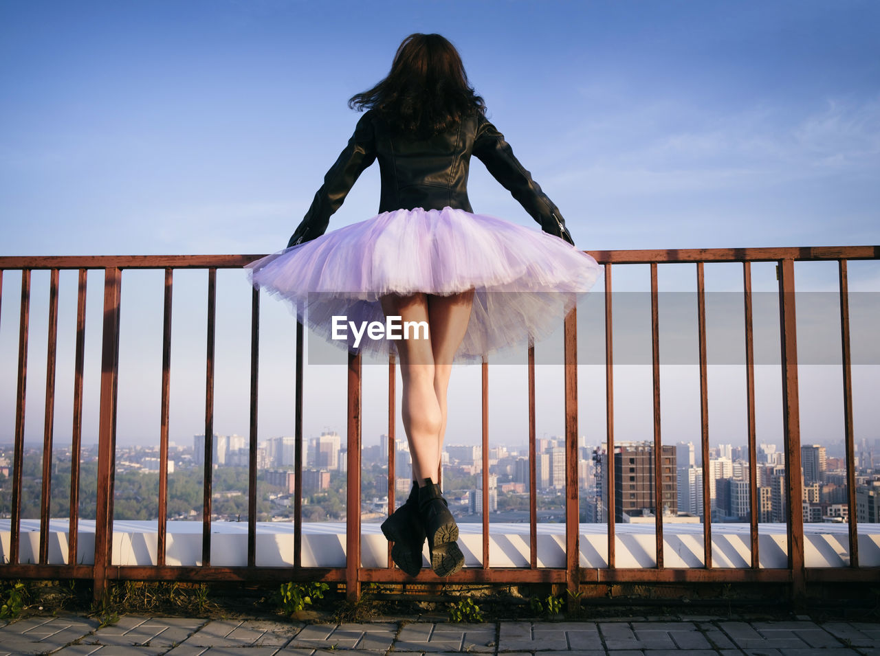 A ballerina in a tutu  jumps holding on to the fence at sunset admiring the panorama of the city