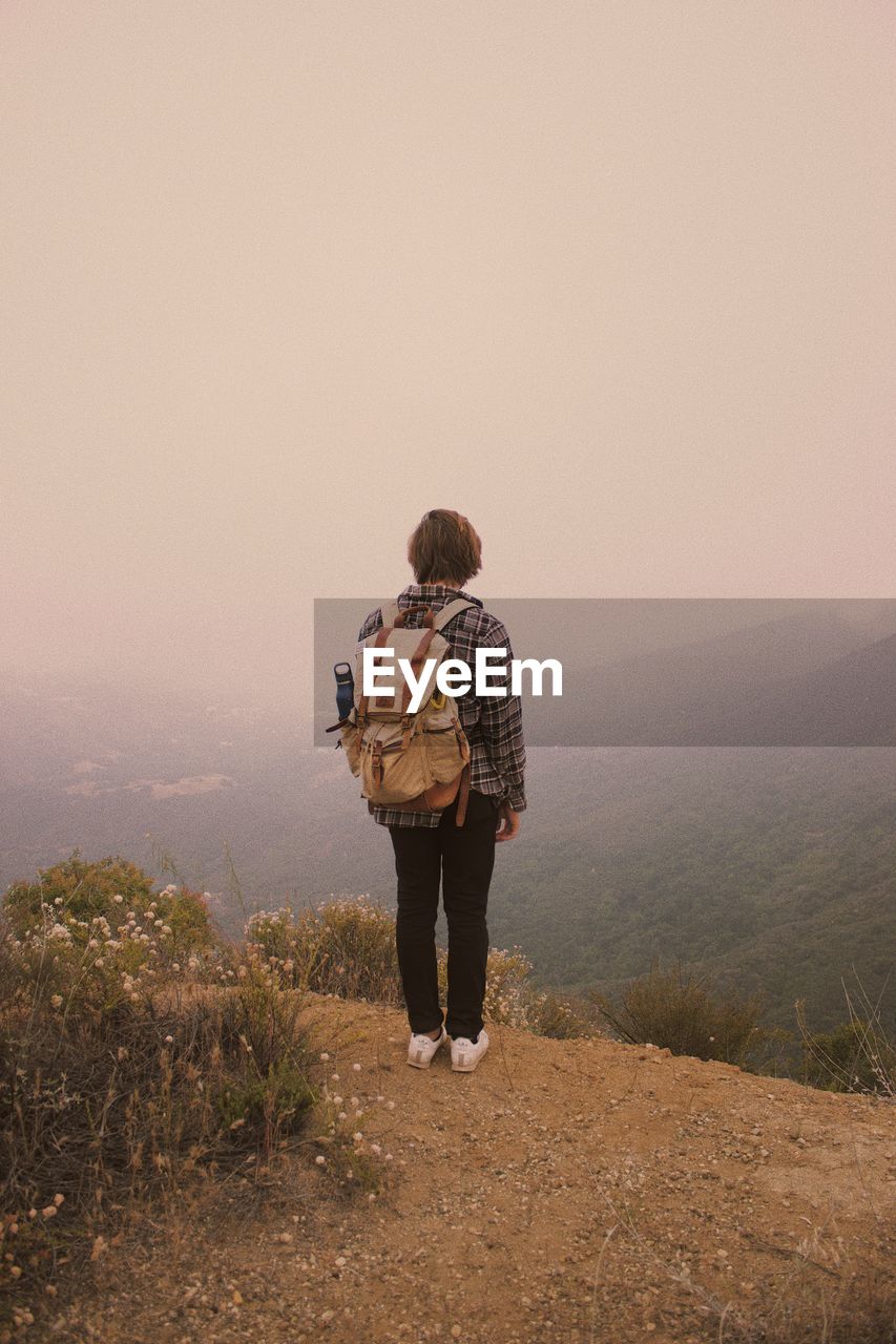 REAR VIEW OF MAN STANDING AGAINST MOUNTAIN