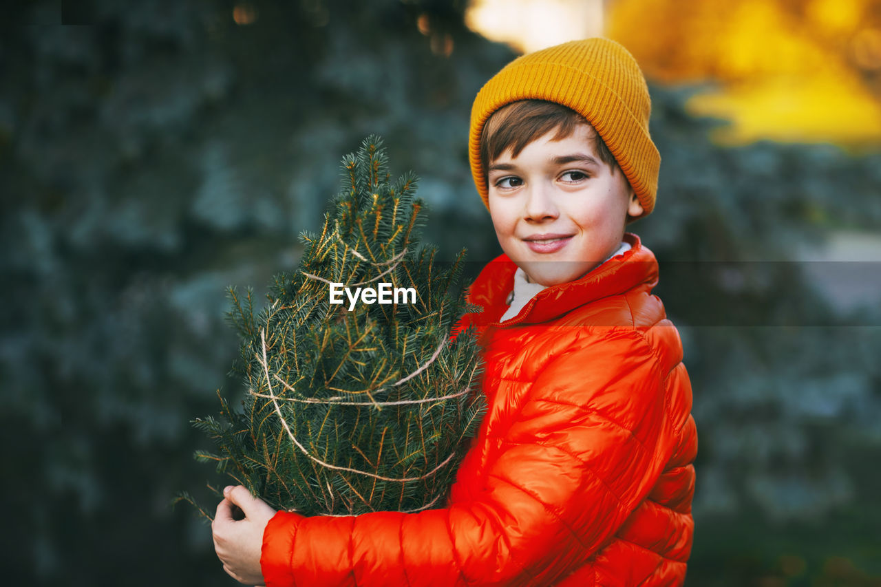 A boy in a bright orange jacket and yellow hat holds a purchased christmas tree in his hands, 