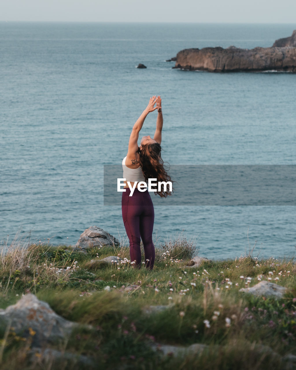 Back view of young fit female with long wavy hair in sportswear performing mountain with arms up and backbend asana while practicing yoga on grassy hill at seaside