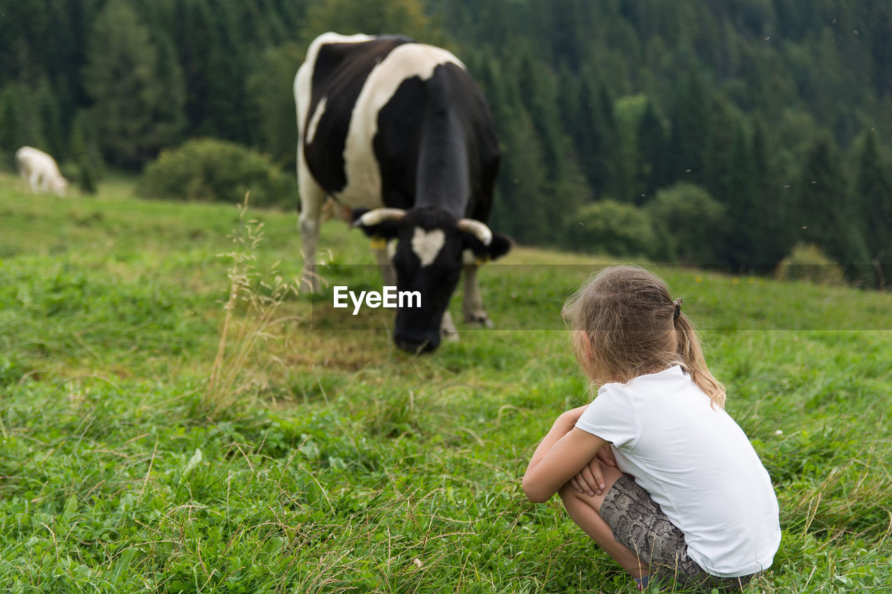 Girl crouching on grassy field with cow in background