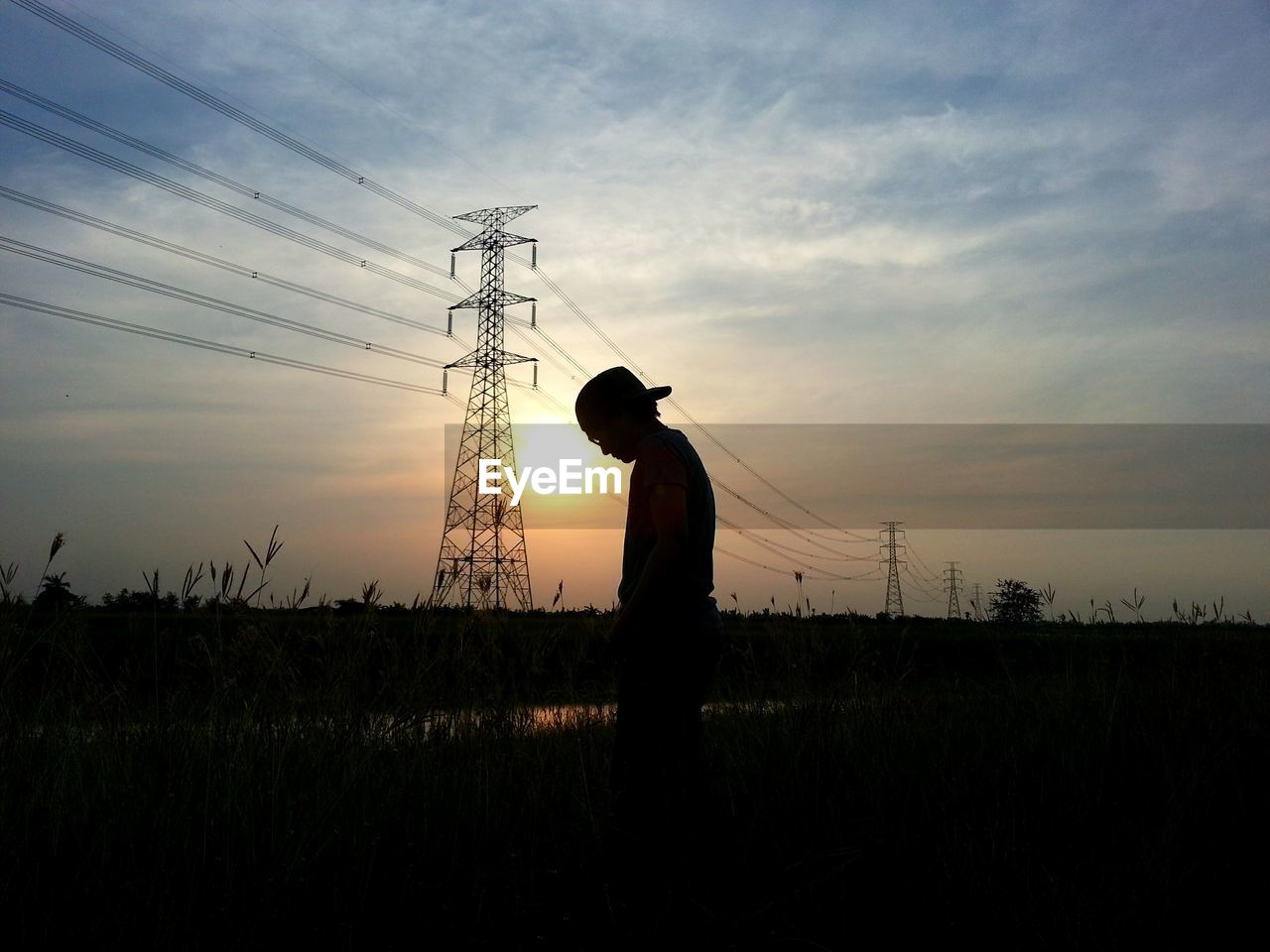 Silhouette man standing on field against electricity pylon in background against sky during sunset