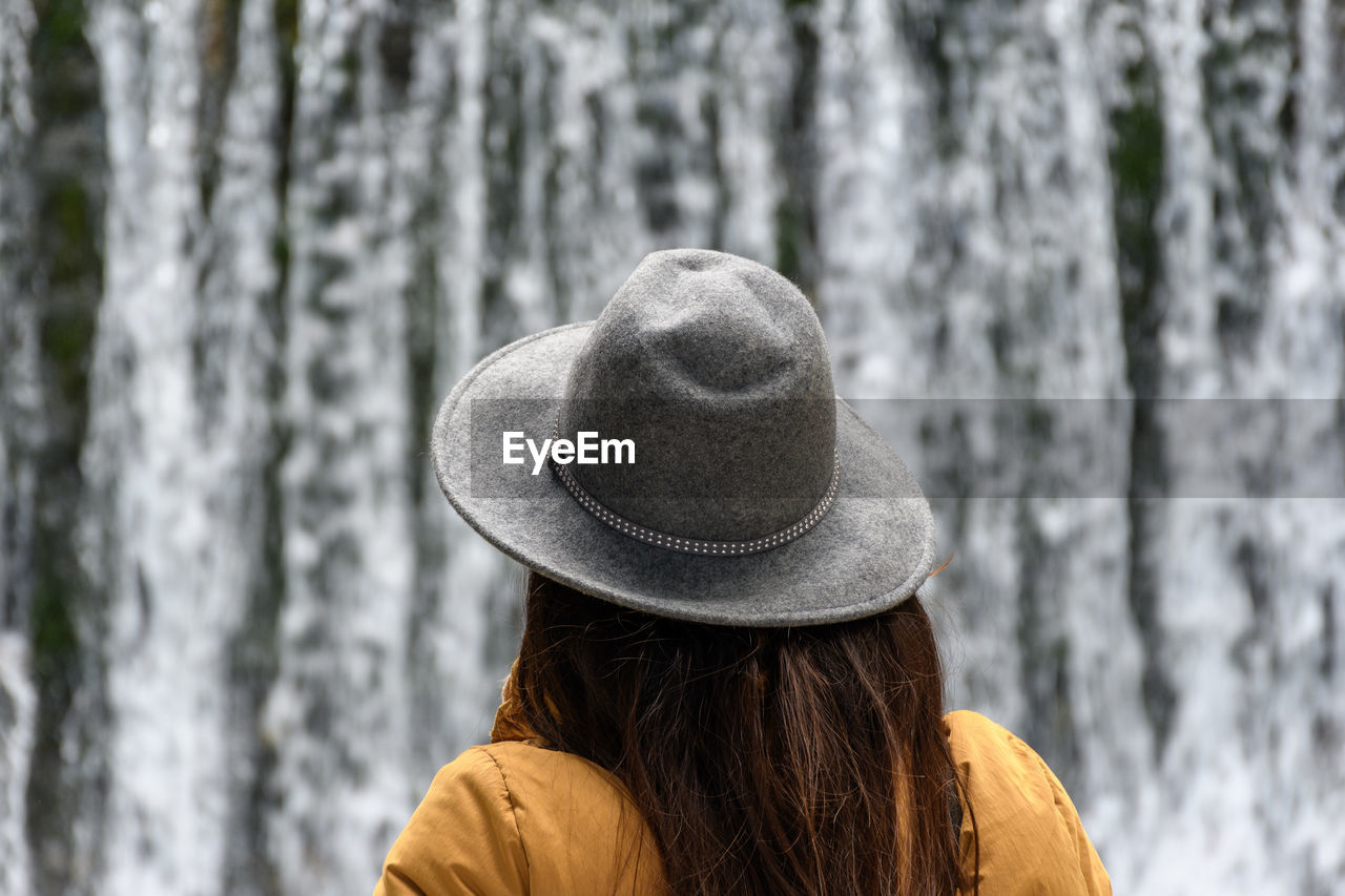 Rear view of woman wearing warm clothes and hat, standing in front of waterfall.