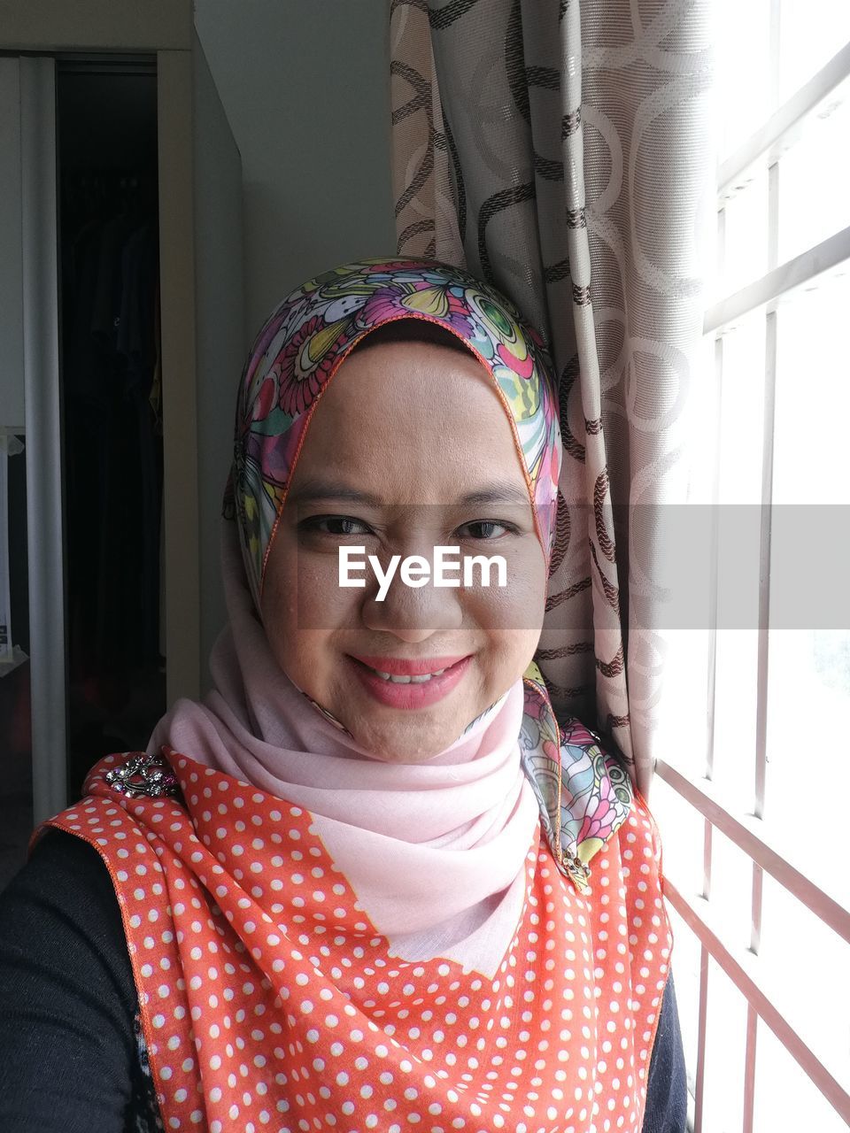 Portrait of smiling woman in hijab by window at home