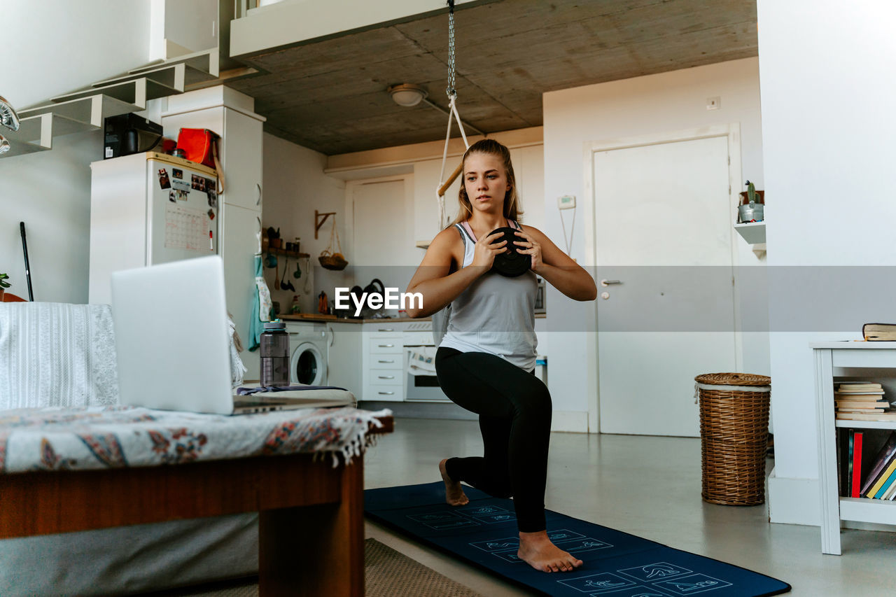 Concentrated young female in sportswear training with weight plate in kitchen at home watching tutorial on laptop