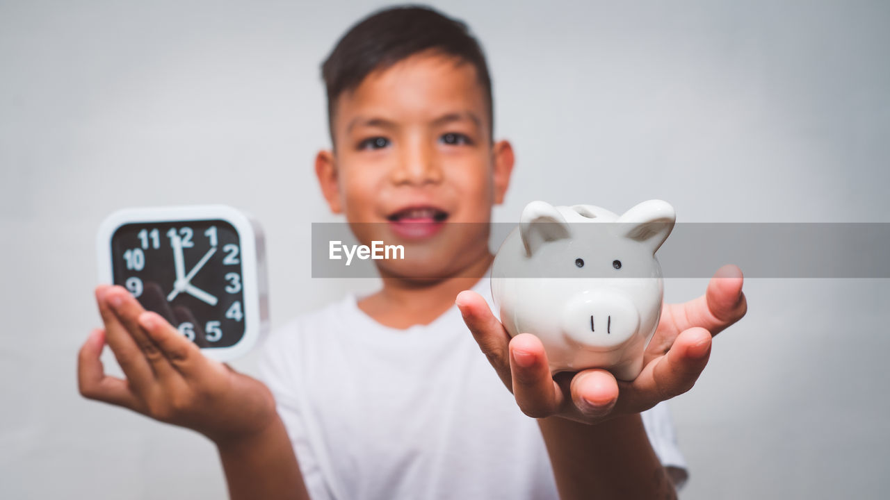 Portrait of boy holding clock and piggy bank against white background