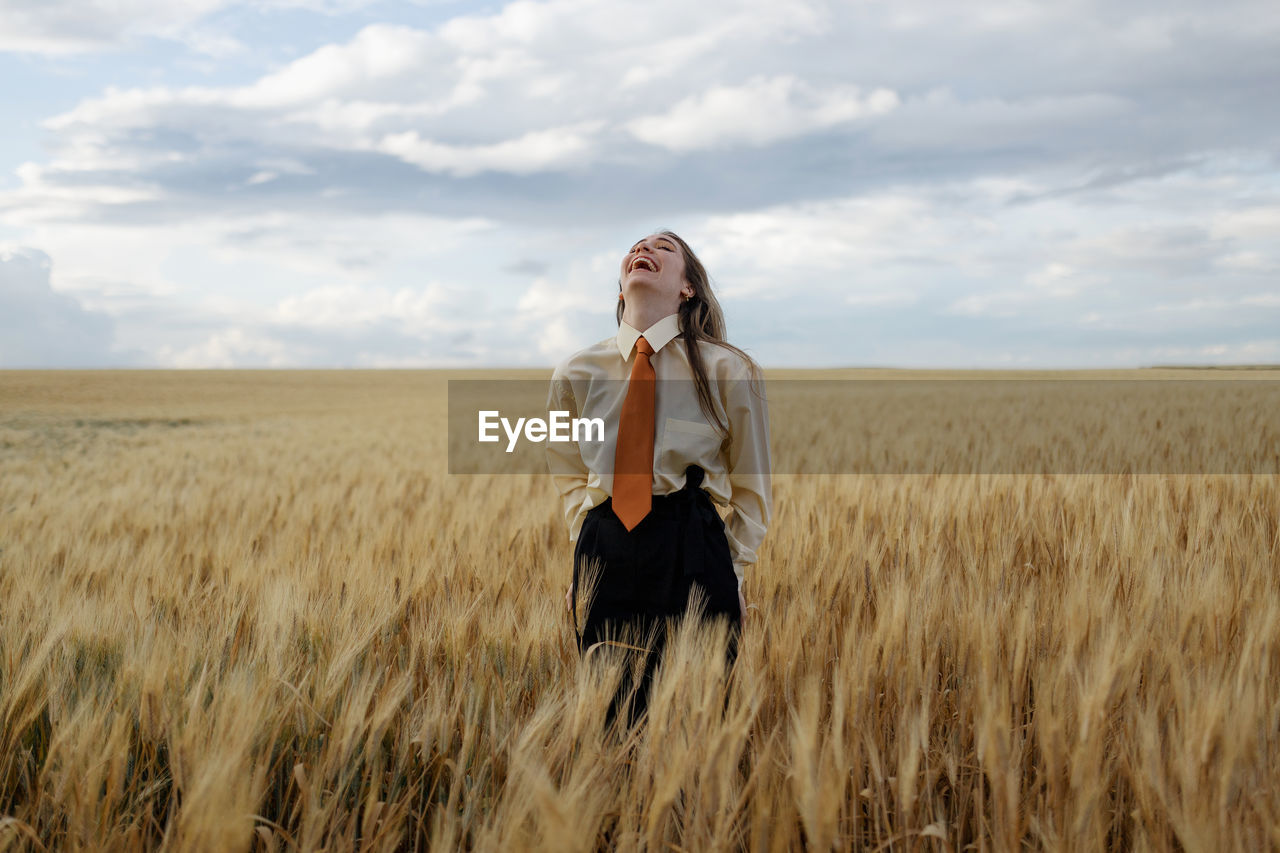 Positive young female in white shirt and red tie standing with hands behind back among wheat spikes in countryside