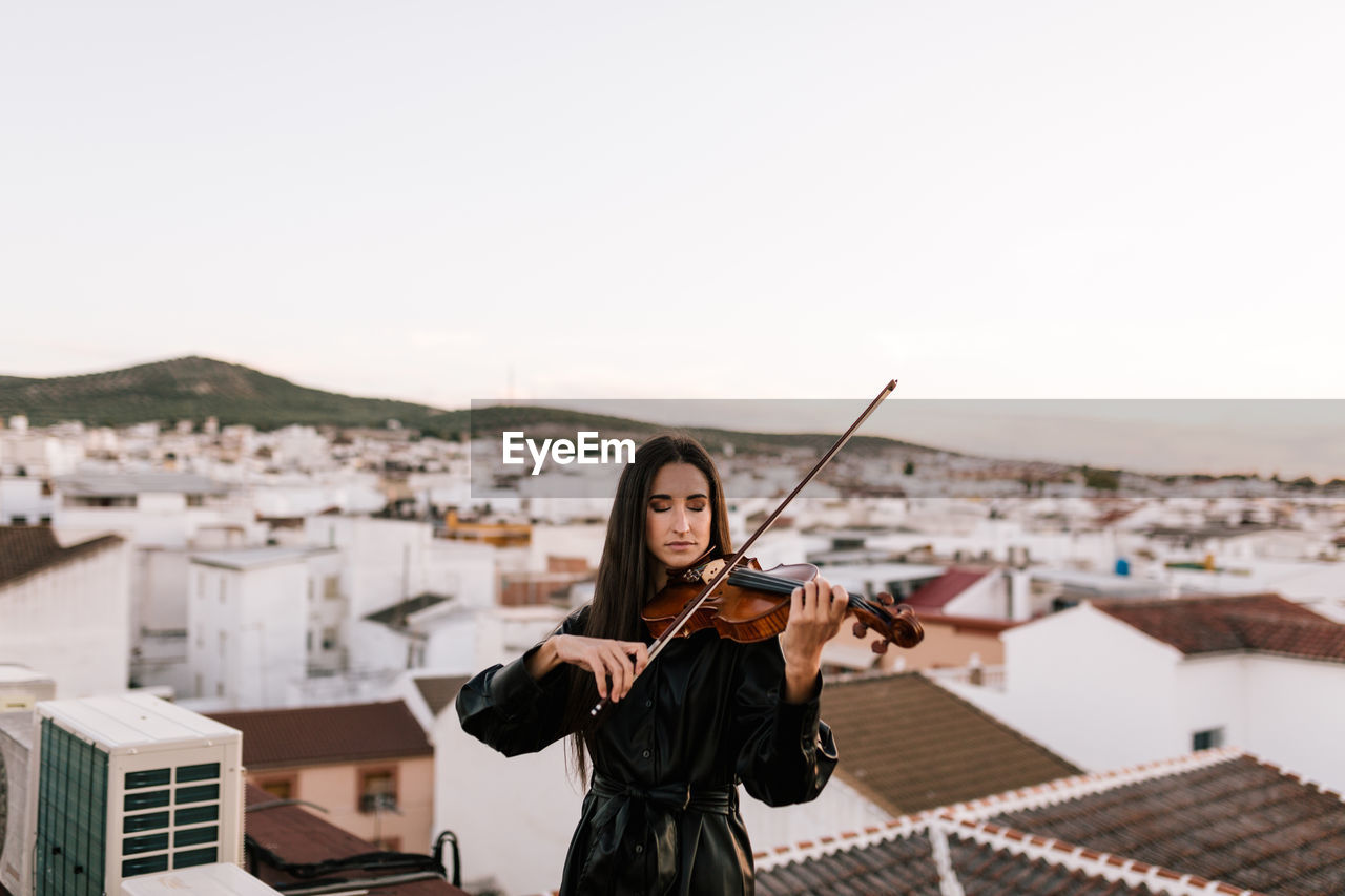 Young beautiful female musician in stylish mini dress holding acoustic violin and standing on rooftop in residential suburb and closed eyes on sunny evening