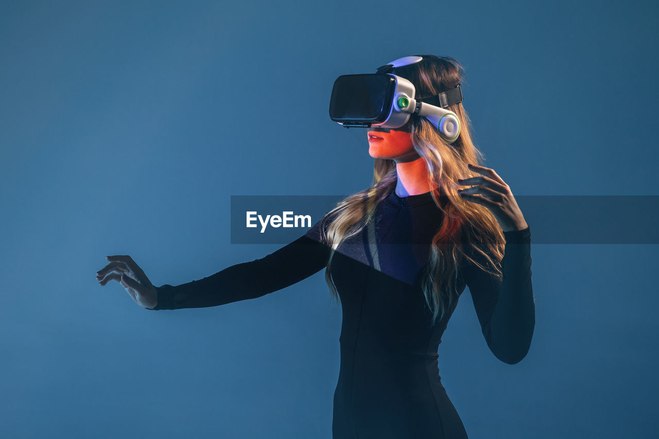 Side view of young unrecognizable blonde woman standing touching air and wearing virtual reality headset on colorful blue studio background