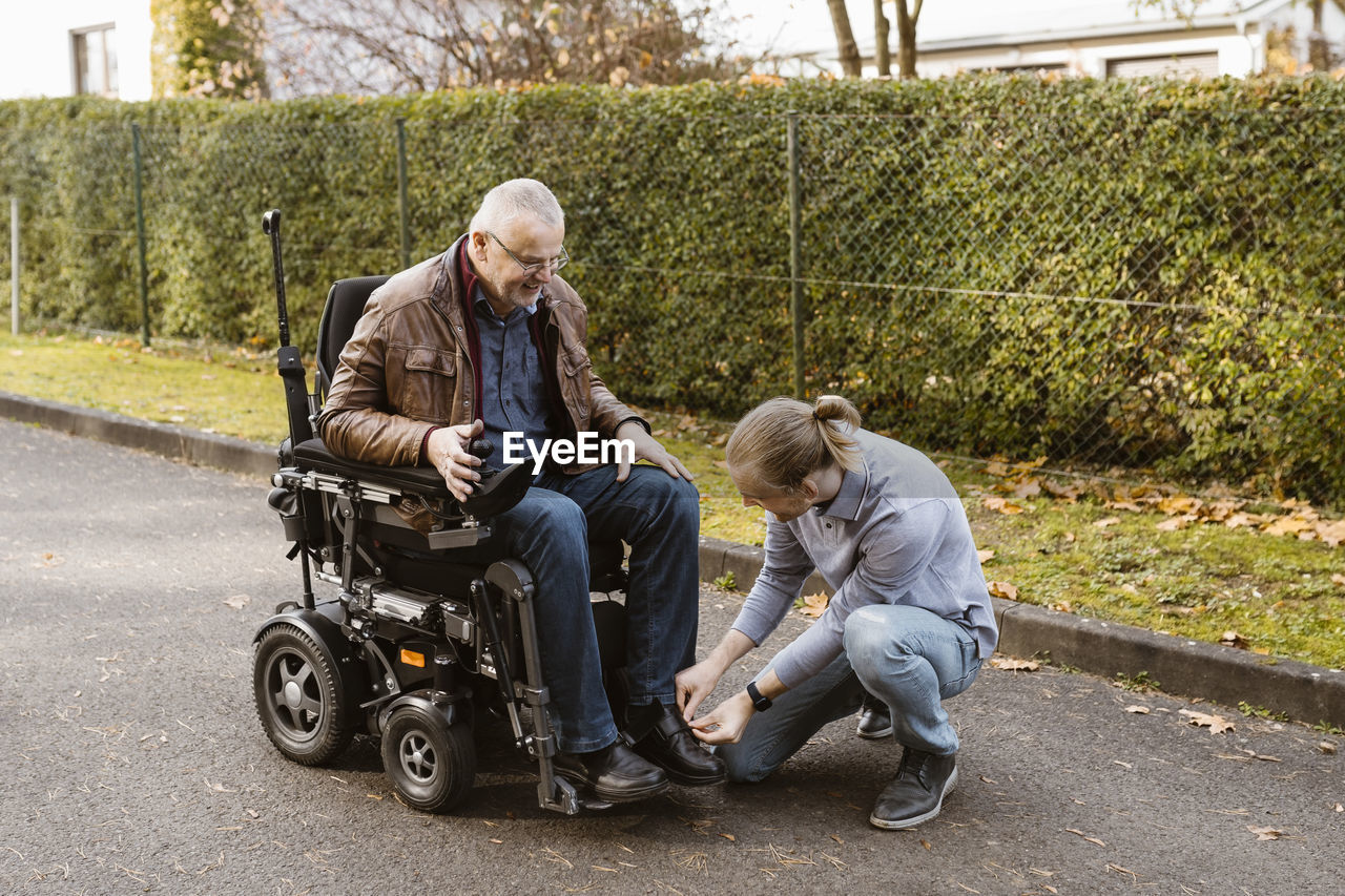 Young man kneeling while tying shoelace of father with disability sitting in motorized wheelchair