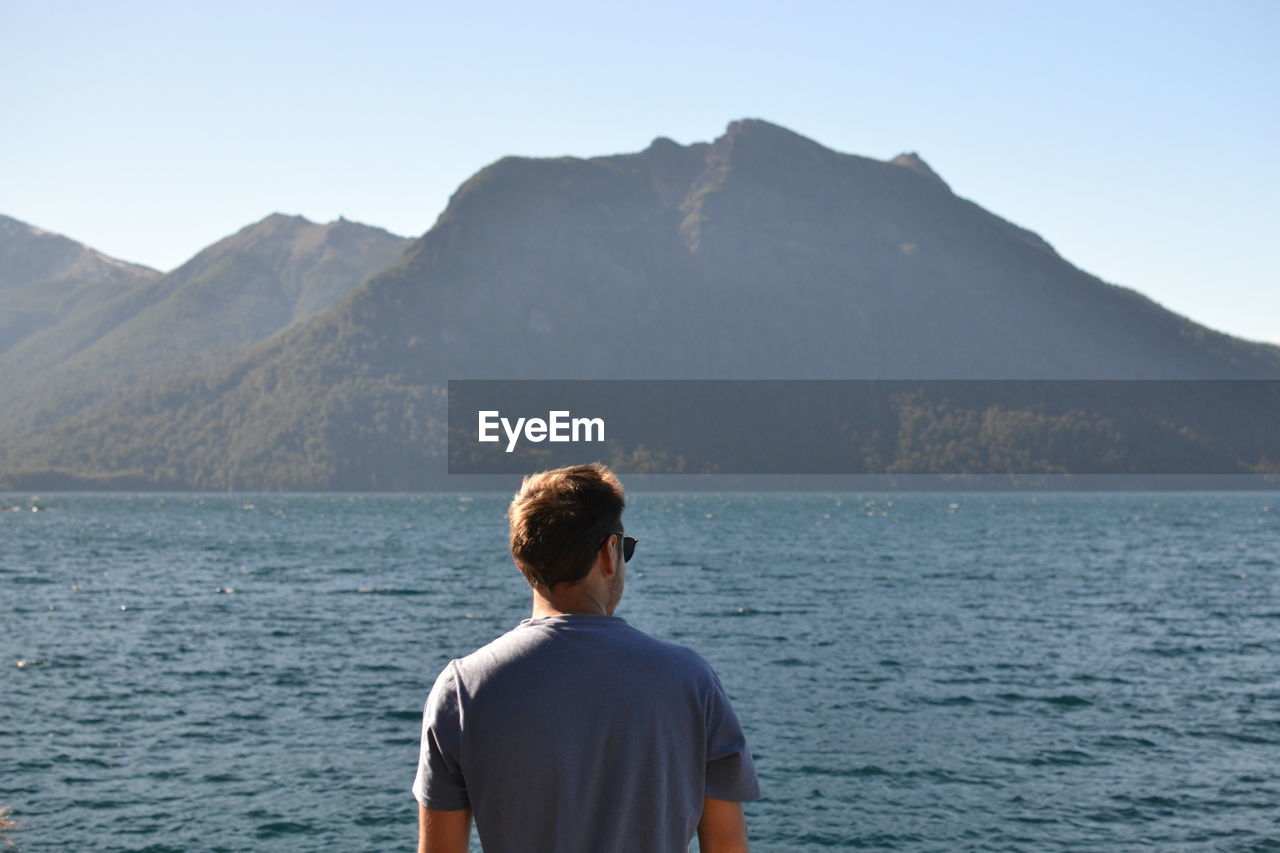 Rear view of man looking at lake and mountains against clear sky