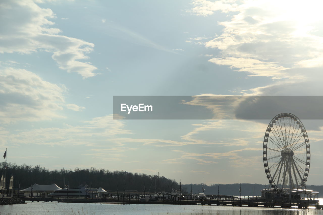 Ferris wheel amidst river against cloudy sky