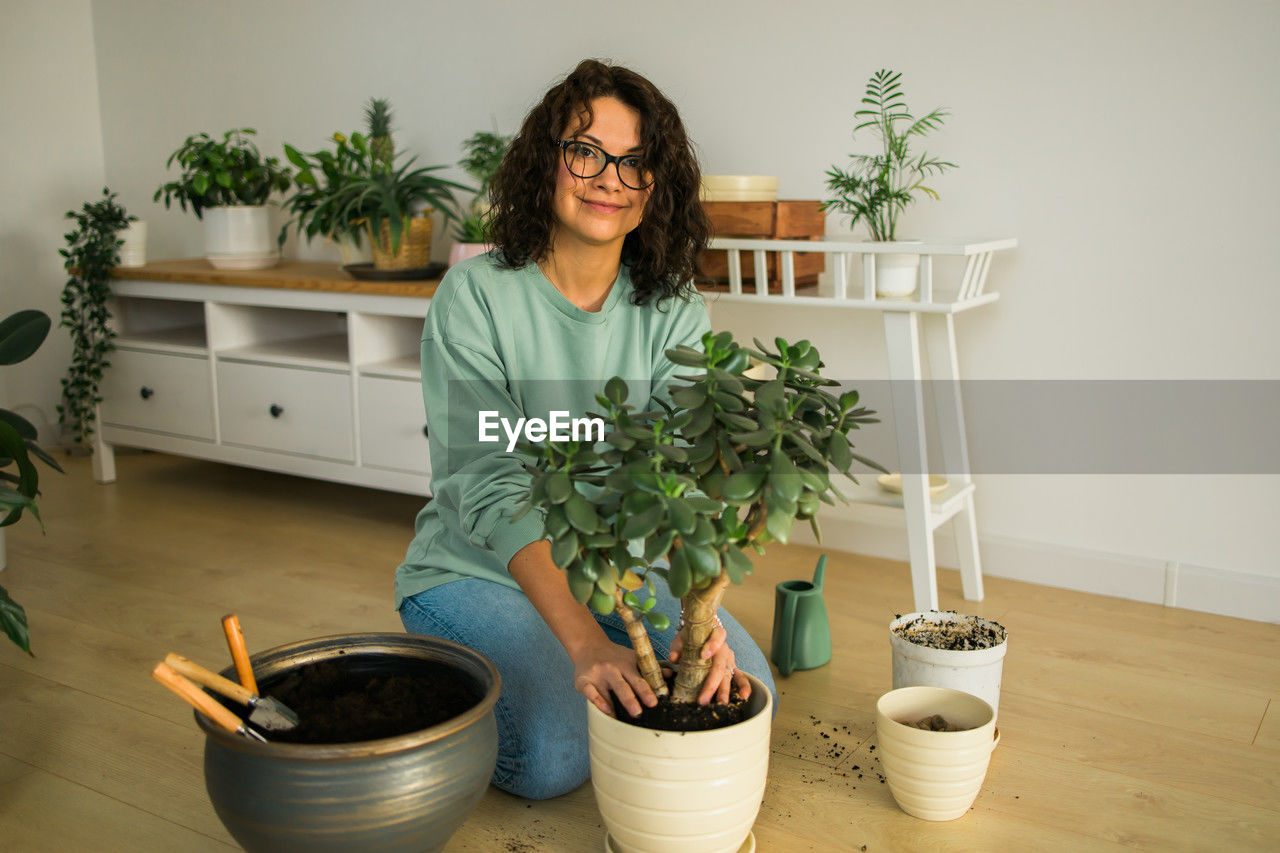 potted plant, one person, adult, plant, indoors, lifestyles, houseplant, flowerpot, growth, domestic life, women, nature, front view, smiling, portrait, young adult, home interior, casual clothing, looking at camera, happiness, holding, hairstyle, glasses, brown hair, domestic room, eyeglasses, female, gardening, herb, emotion, food and drink, standing, table, food, floristry