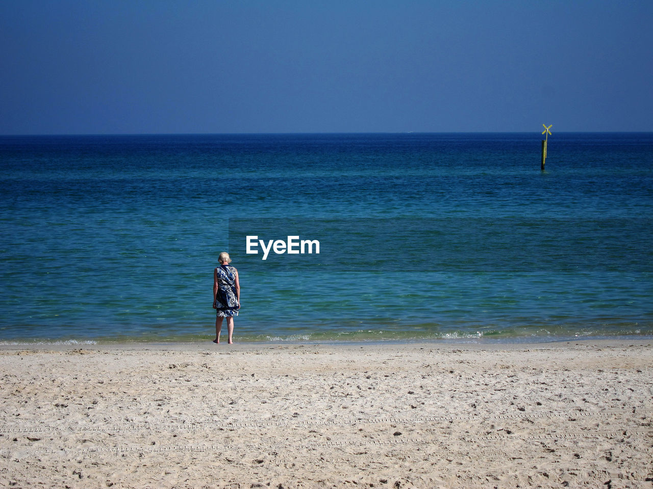 Rear view of woman standing at beach against clear blue sky during sunny day