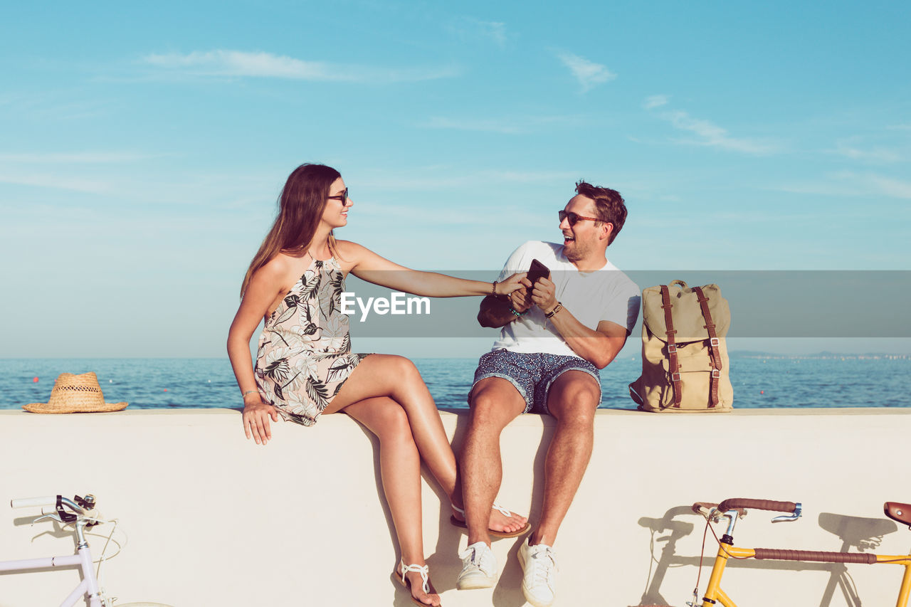 Couple enjoying while sitting on retaining wall by sea against sky