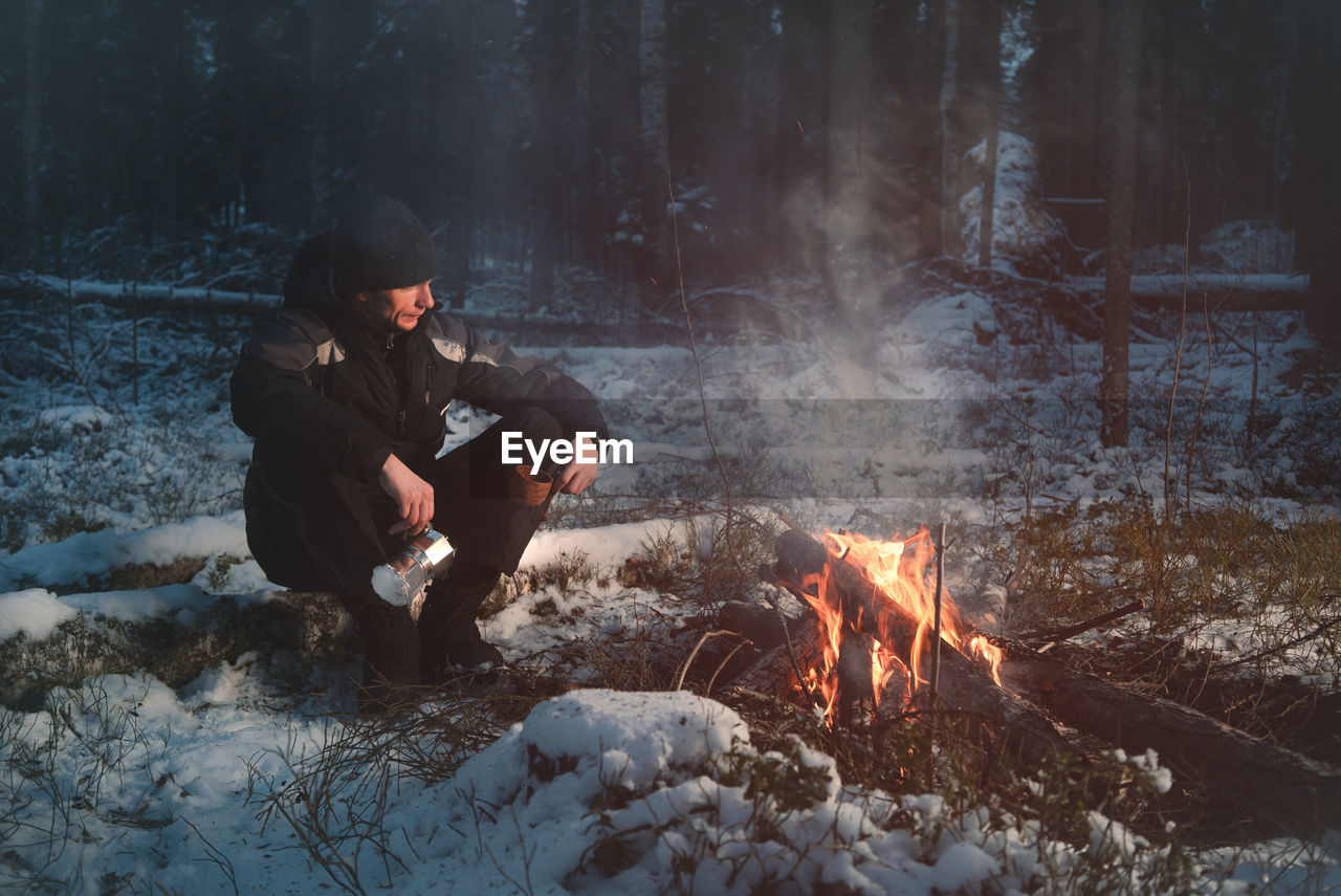 Full length of man sitting by campfire in forest during winter