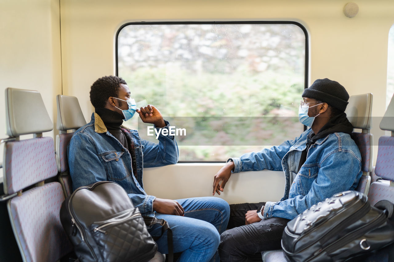 Side view of calm black men in medical masks sitting on passenger seats and looking out of window while traveling by train during covid 19 epidemic