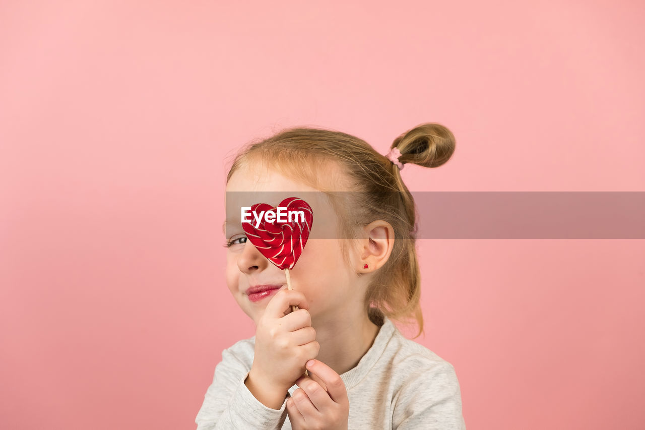 Cute girl holding heart shape on eye against colored background