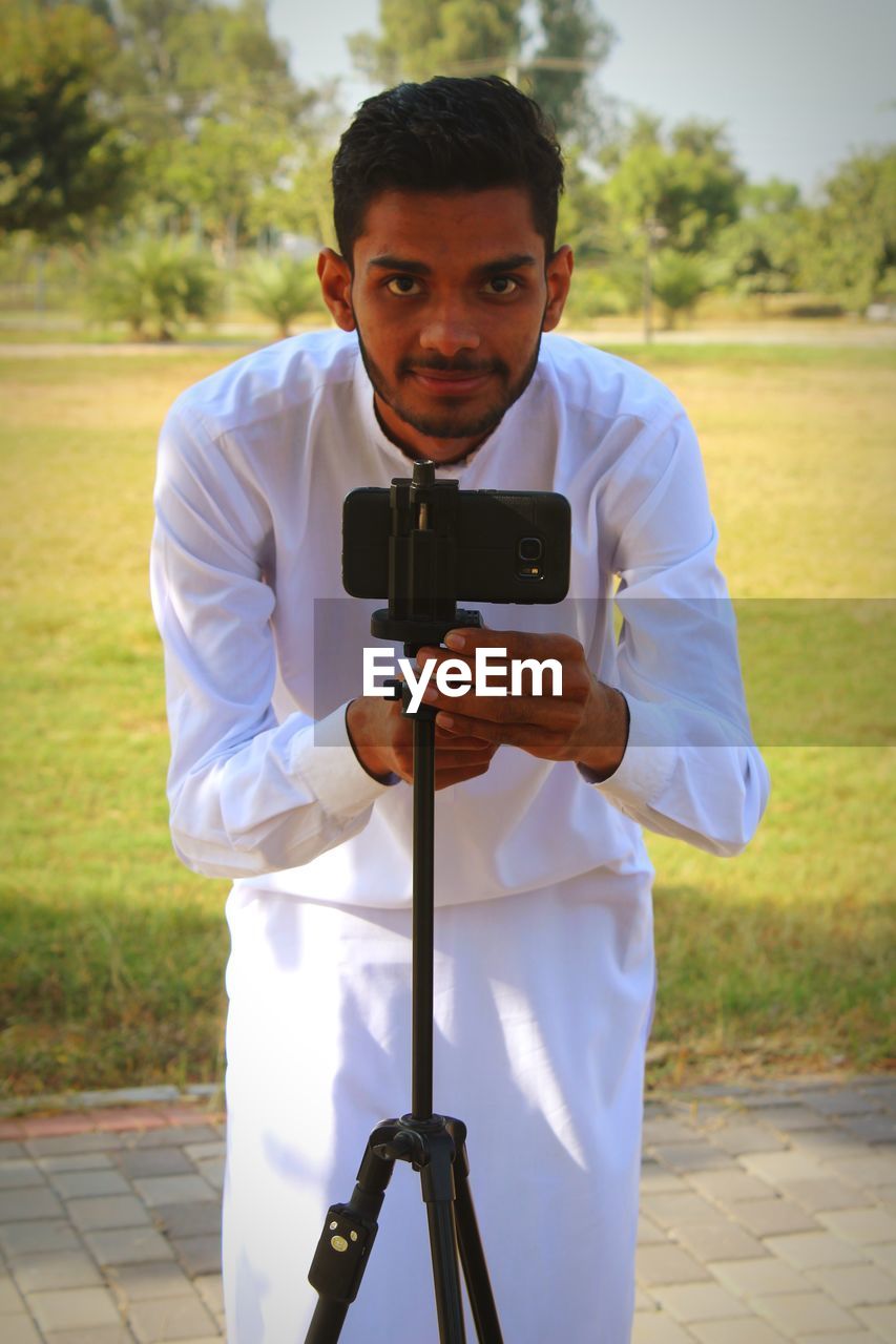 one person, technology, adult, portrait, men, standing, looking at camera, activity, young adult, holding, person, tripod, wireless technology, communication, front view, occupation, spring, clothing, camera, outdoors, leisure activity, lifestyles, day, nature, smiling