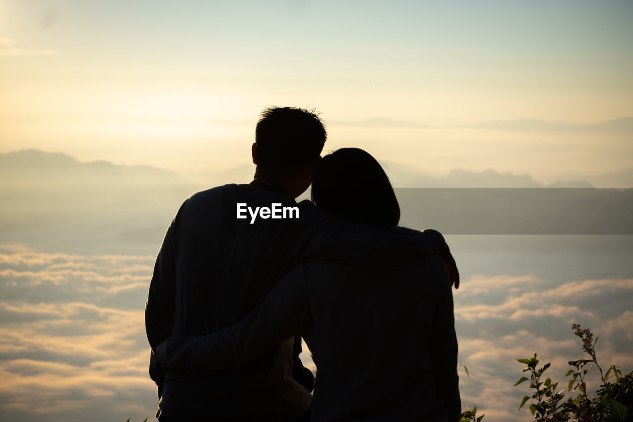 Rear view of couple with arm around sitting against sky during sunset