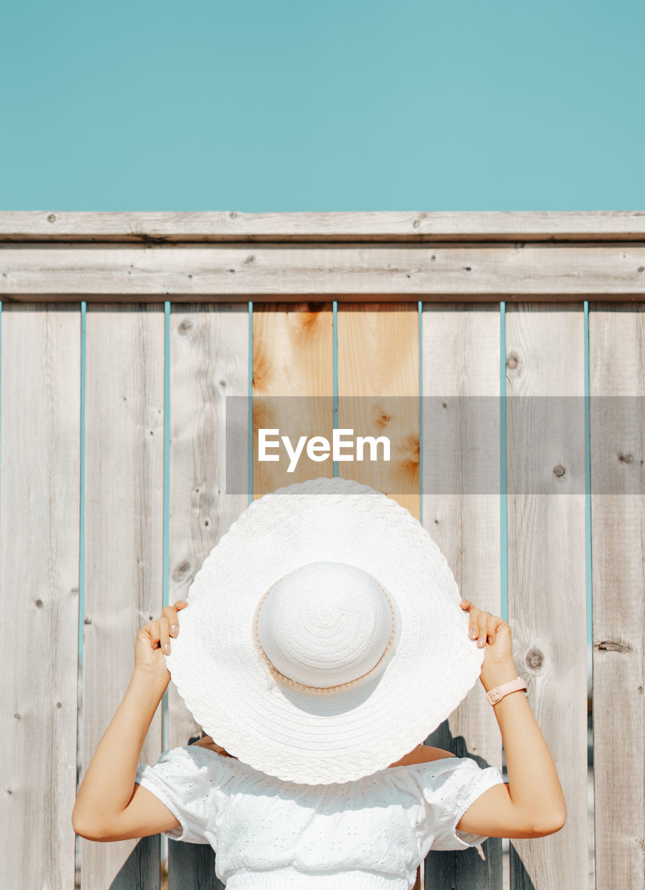 Young woman covering face with hat against wooden wall outdoors