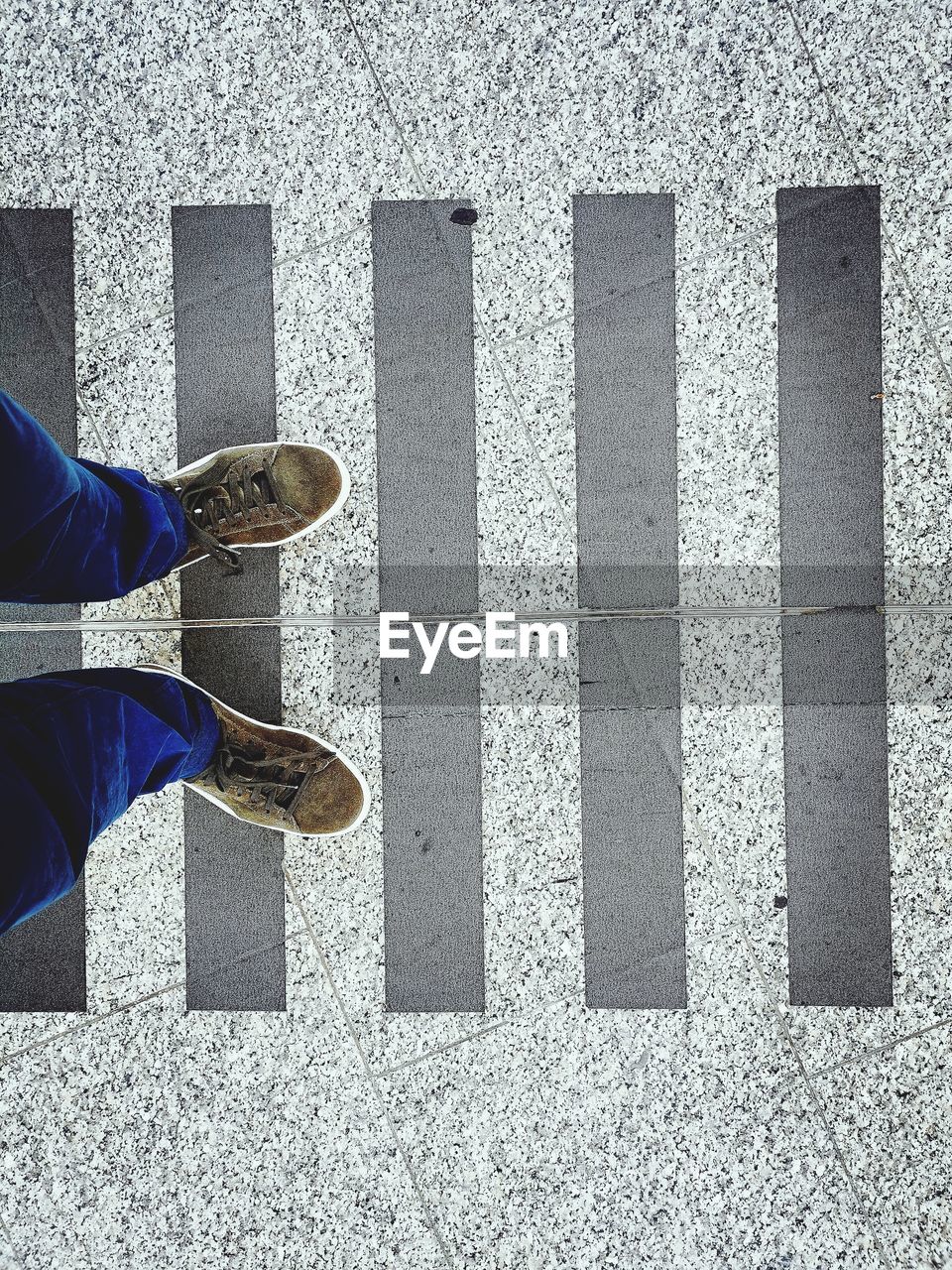 low section, human leg, one person, shoe, high angle view, standing, day, lifestyles, footwear, human limb, men, human foot, leisure activity, directly above, wall, city, flooring, limb, blue, street, outdoors, personal perspective, black, sunlight, pattern, road, casual clothing, floor, transportation, adult, sign, white, asphalt, shadow