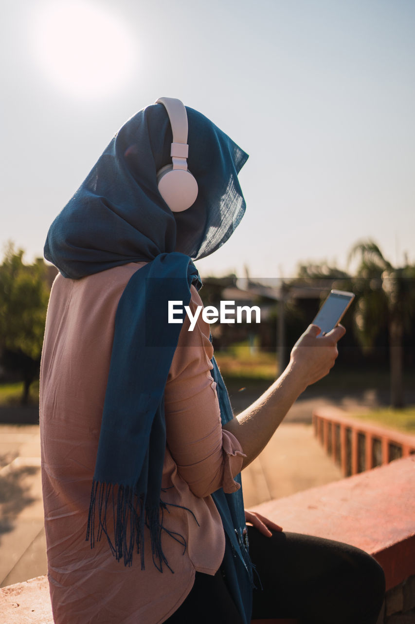 Side view of arab female in headscarf and headphones browsing mobile phone and listening to music while enjoying sunny day in city