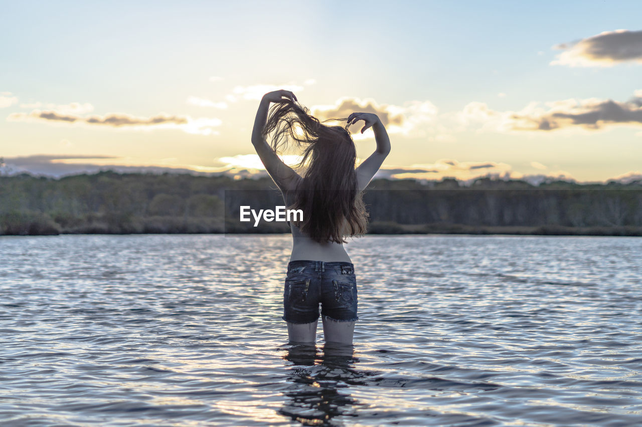 Rear view of shirtless woman standing in lake against sky