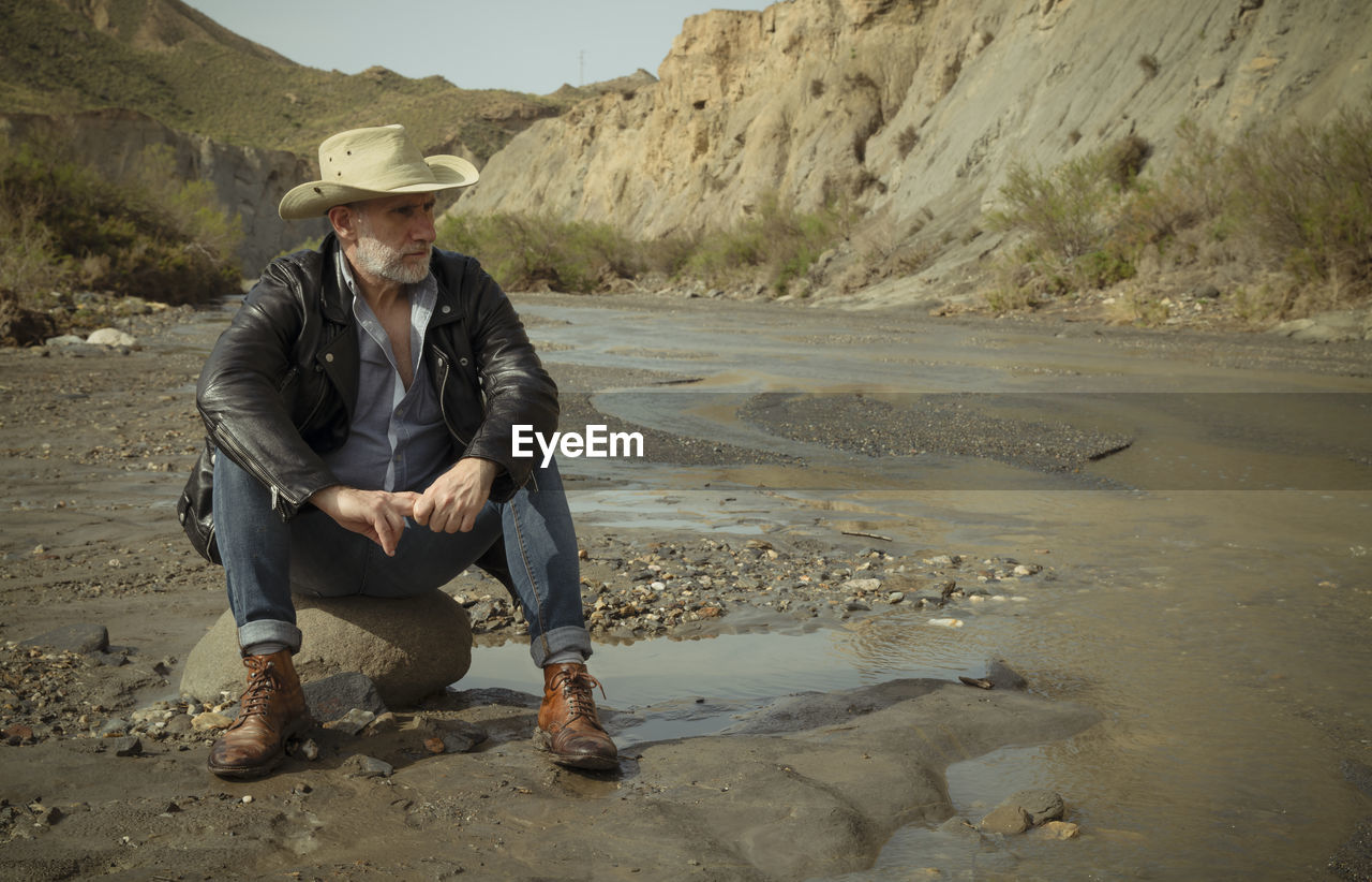 Adult man in cowboy hat sitting on rock along river flows in desert. almeria, spain